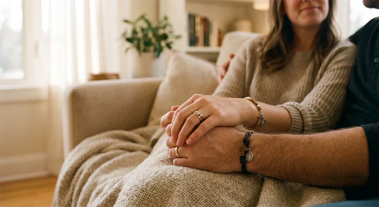 A close-up shot captures a couple’s hands clasped tightly together in soft light, showcasing their wedding rings and a delicate bracelet featuring a cross—a symbol of faith—on the wrist.